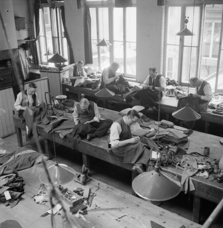 Savile Row - Tailoring at Henry Poole and Co., London, England, UK, 1944. Copyright IWM Non-commercial license.  A view of the workroom at Henry Poole and Co., showing tailors at work on various types of jacket, including a naval officer's jacket, second from right on the rear row. The men are all sitting on the workbenches, some cross-legged, the garments resting in their laps as they work.