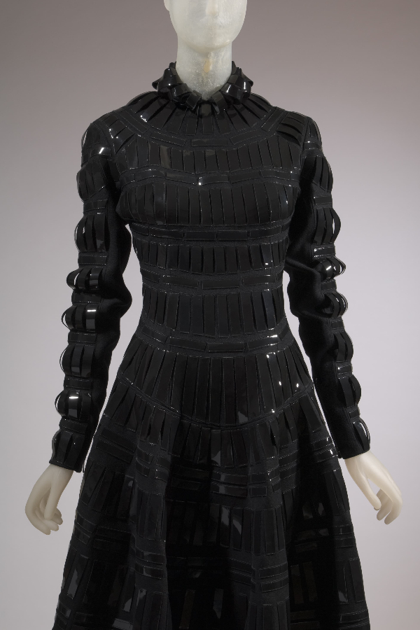 High neckline, long sleeve, full skirt black wool dress. Dress has vertical strips of patent leather from neckline to hem and from shoulder to wrist.