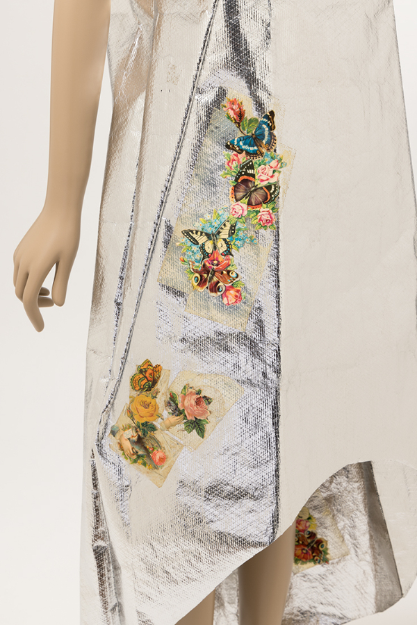 Skirt of metallic silver Mylar tent-silhouette dress with high-low hem, applied paper flower and butterfly motifs along the wearer’s right side and another at the back inside hem.