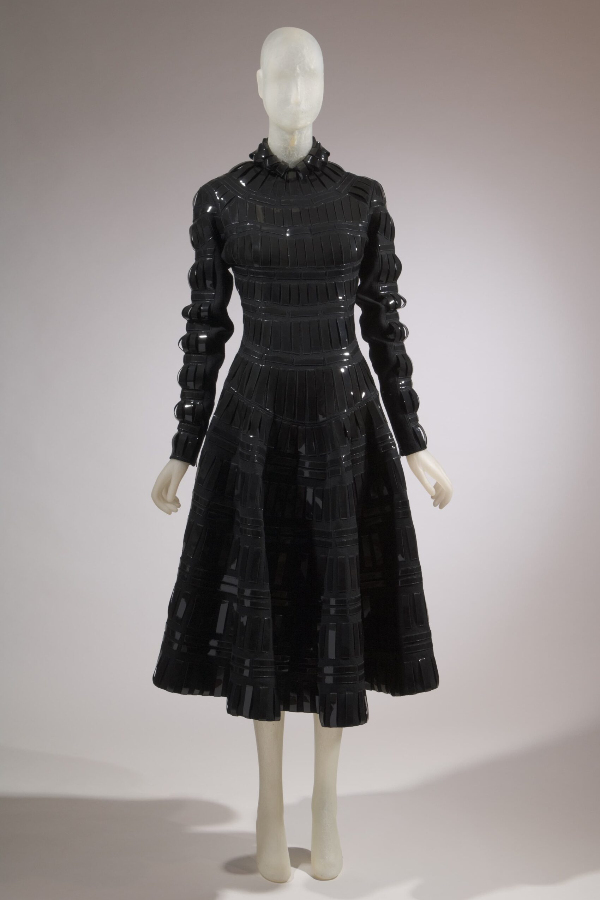High neckline, long sleeve, full skirt black wool dress. Dress has vertical strips of patent leather from neckline to hem and from shoulder to wrist.