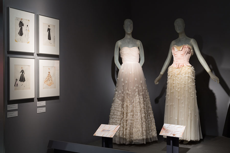 On wall: Sketches, Fashion Institute of Technology|SUNY, FIT Library Special Collections and FIT Archives Left: Licensed copy of Pierre Balmain’s Angel evening dress, created by Orcilia, tulle, feathers, 1946-1947, Cuba, 80.304.1, gift of Molly Milbank Right: Bergdorf Goodman Custom Salon, licensed copy of a Pierre Balmain evening dress, silk satin, coq feathers, 1949, USA, 91.244.1, gift of Barbara Louis Each season, buyers from Bergdorf Goodman traveled to Paris to purchase 80-100 samples from various couture houses. Bergdorf’s was known for creating the highest quality copies by using the same couture fabrics as the original and fitting each client three times to ensure a proper fit. The quality of Bergdorf’s construction is evident in the structured silk satin bodice of this dress.
