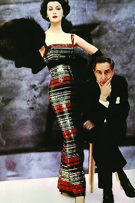 photograph of Dovima wearing a red, black, and white Norell dress standing beside a sitting Norman Norell in a black suit