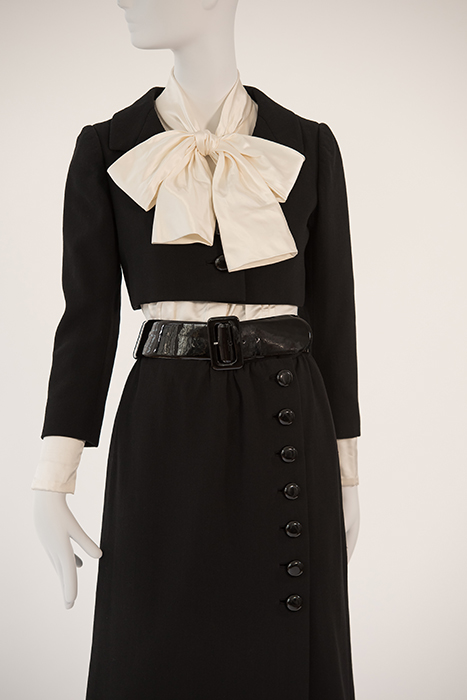 black long sleeve jacket on top of ivory blouse with bow at neck paired with black skirt with black buttons on down right side and black belt at wait