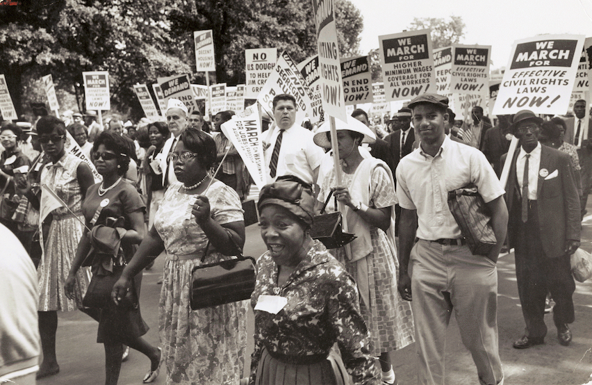 Black and white men and women march on Washington on August 23, 1963 holding signs. One visible sign says We March For Effective Civil Rights Laws Now