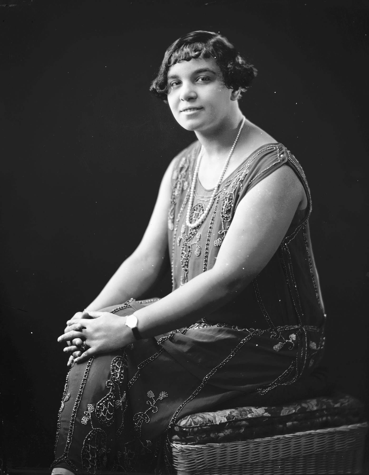 Anges Thomas sit facing the camera with her hand on her knee. She is wearing a flapper style dress with heavy embellishments and a long strand of pearls, circa 1920.