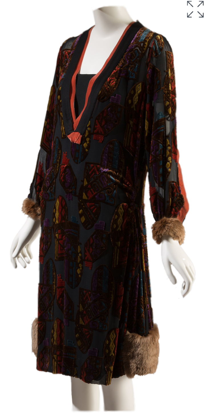 Front view of 1929 'Zanbaraza' motif knee length, long sleeve black chiffon cut velvet dress. The sleeve openings and hem are trimmed with fur. A multicolored shield motif repeats throughout the dress.