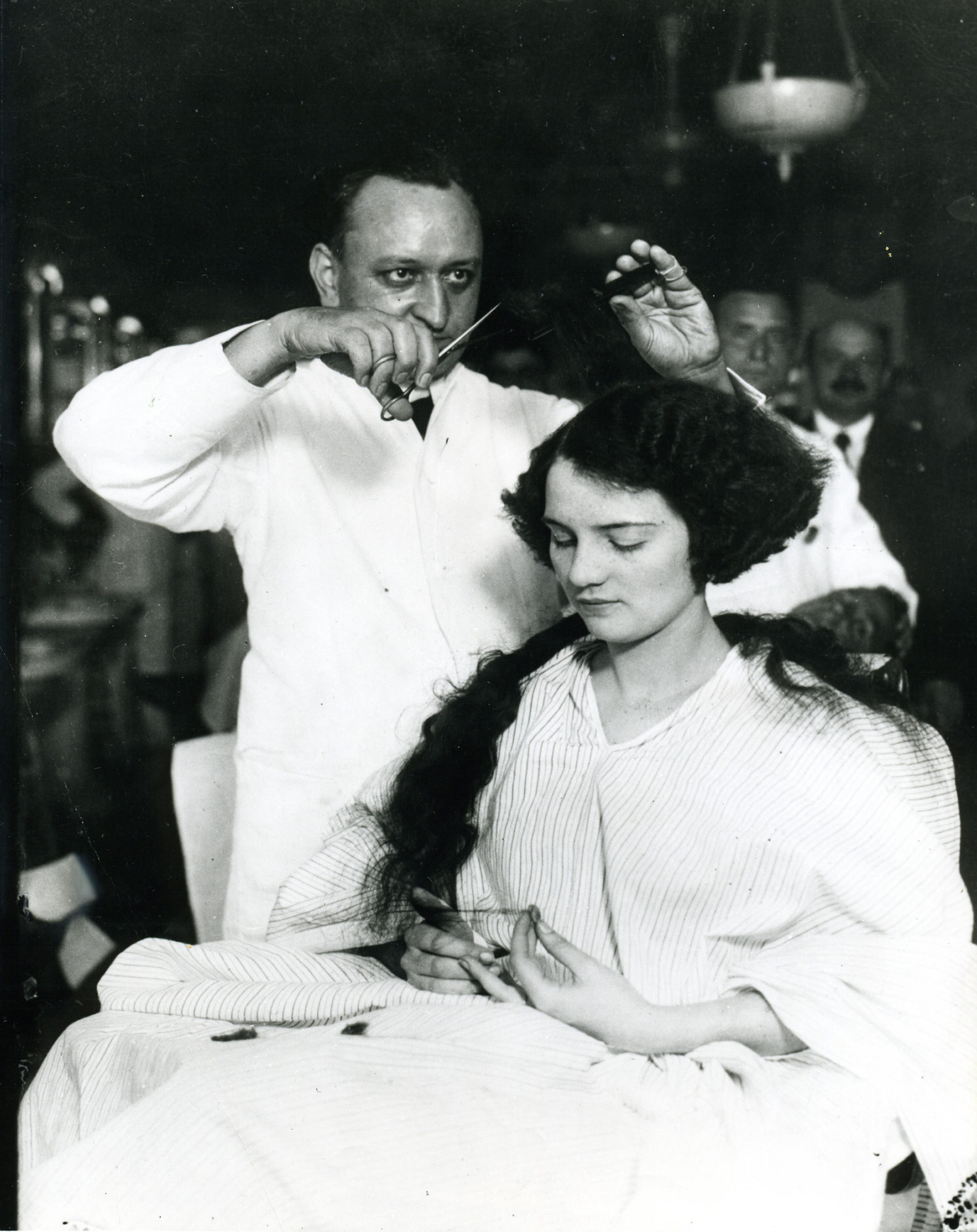 A young woman sits in a chair while a barber bobs her hair, circa 1920