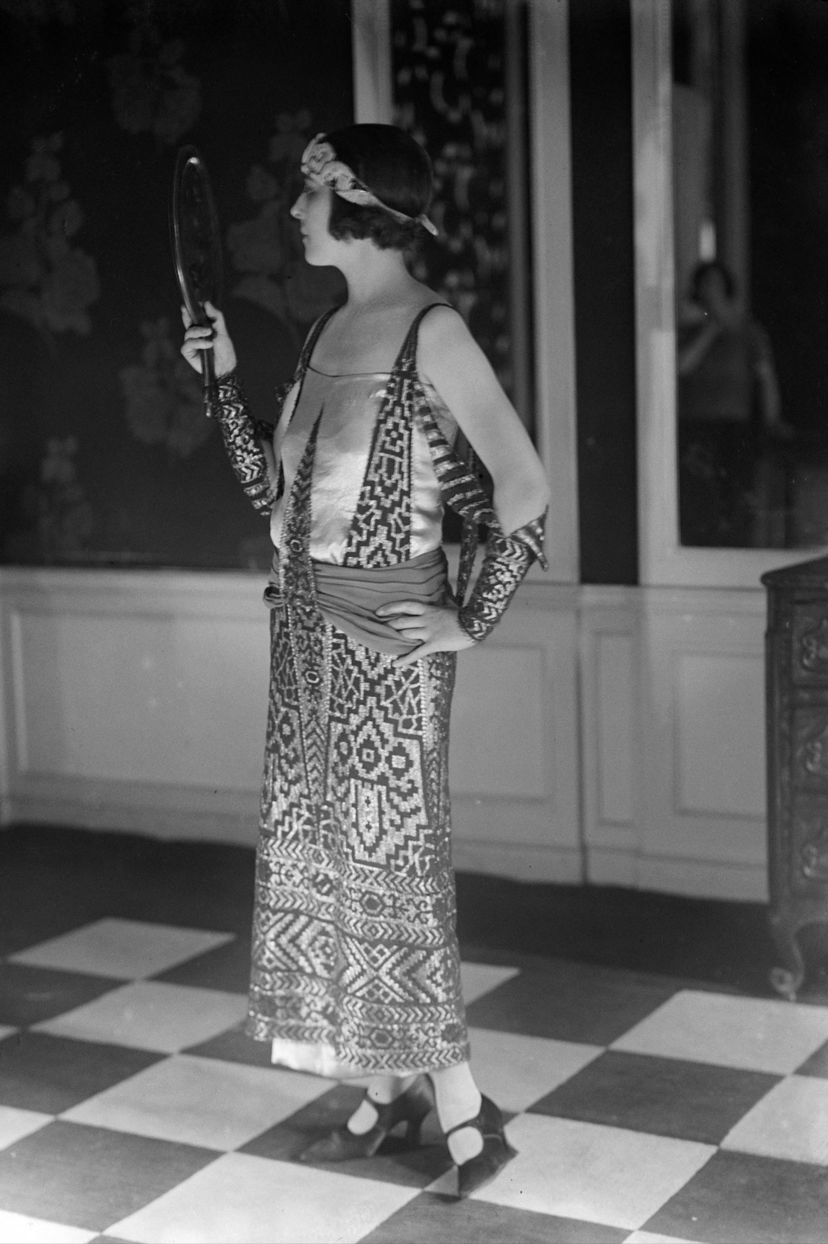 A woman facing away from the camera and looking into a mirror with bobbed hair wearing a ankle length dress with Egyptian-style motifs by Paul Poiret, 1923