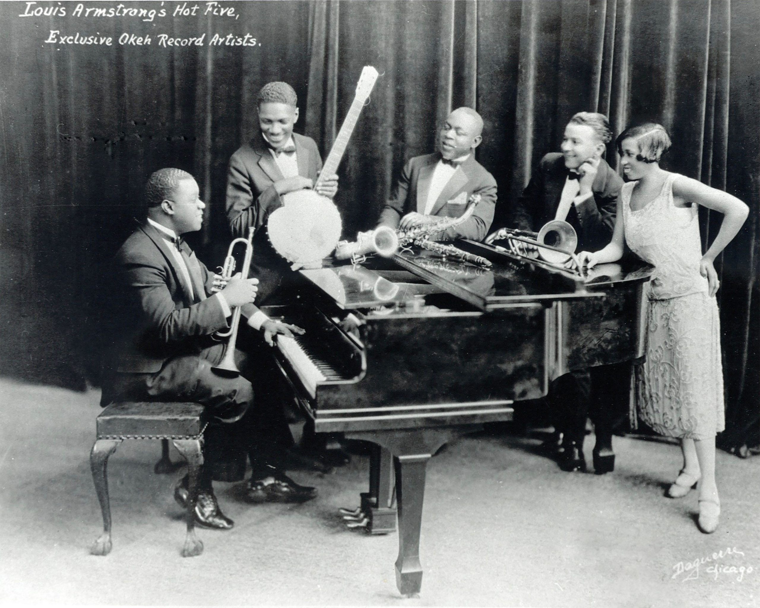The original members of Louis Amrstrong's Hot Five Band sit and stand around a piano in Chicago, IL, 1926