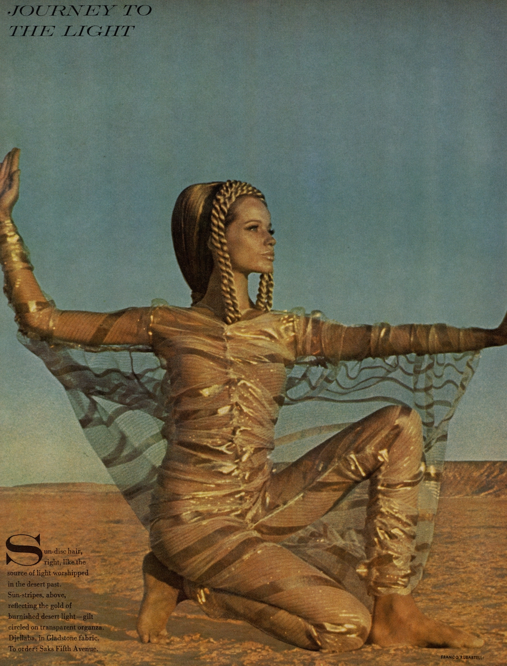 A model poses in the desert in Egypt kneeling on the ground with her arms fully extended out to her sides. She is wearing sheer gold striped organza.