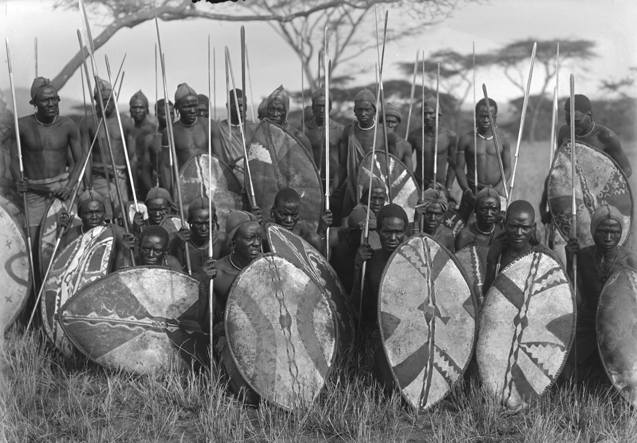 A group of men in Eastern Kenya, some sitting down and some standing, pose holding spears and pigment decorated shields, 1926