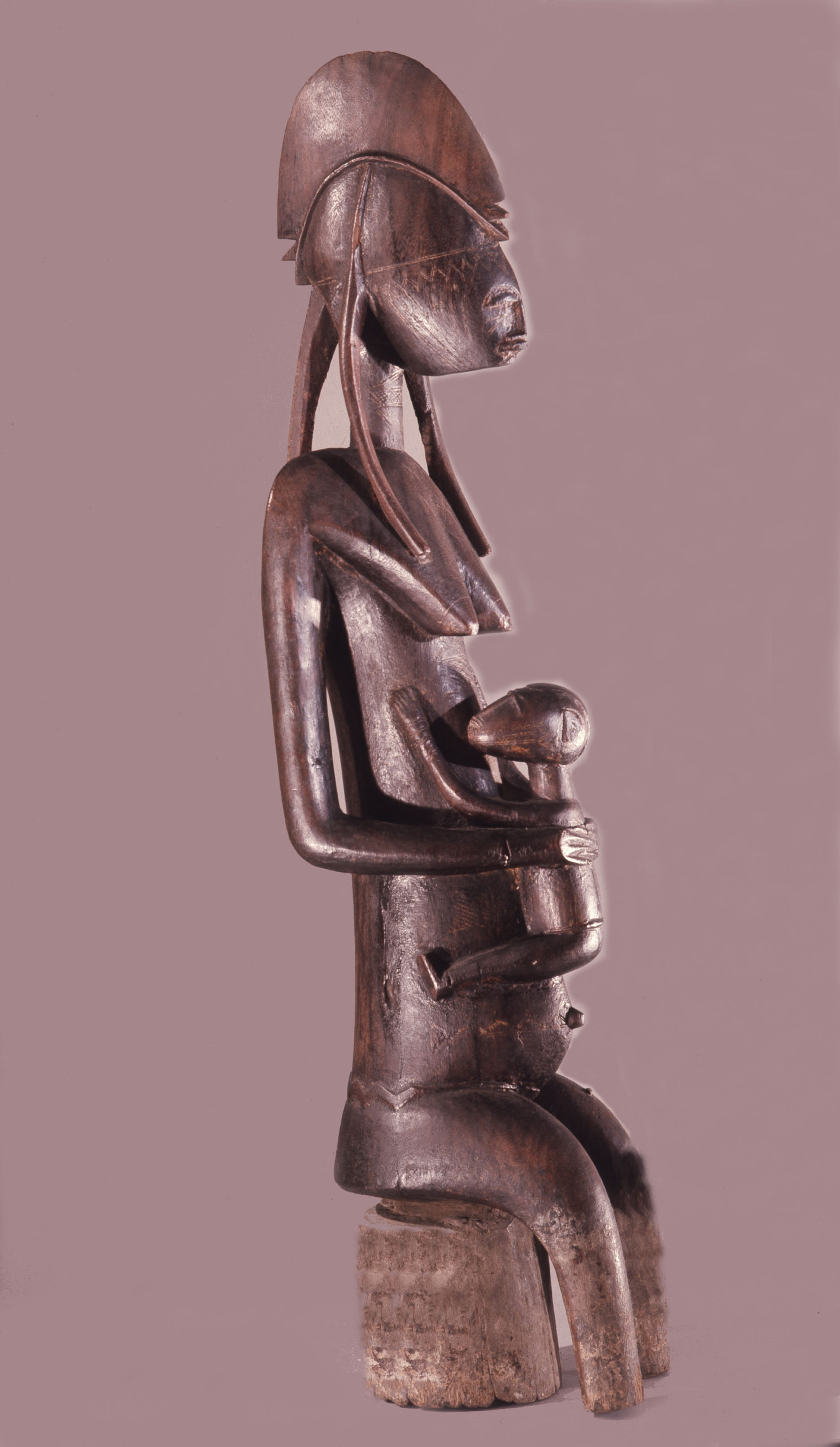 A side view of a wooden sculpture of a seated woman holding a baby with an elongated body and pointed breasts
